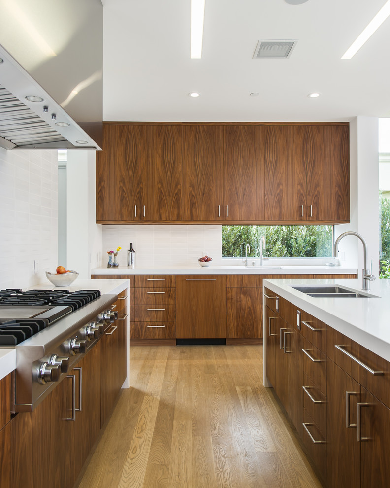 This is an example of a mid-sized modern l-shaped open plan kitchen in Los Angeles with a double-bowl sink, medium hardwood floors, with island, flat-panel cabinets, dark wood cabinets, quartz benchtops, white splashback, ceramic splashback, stainless steel appliances and beige floor.