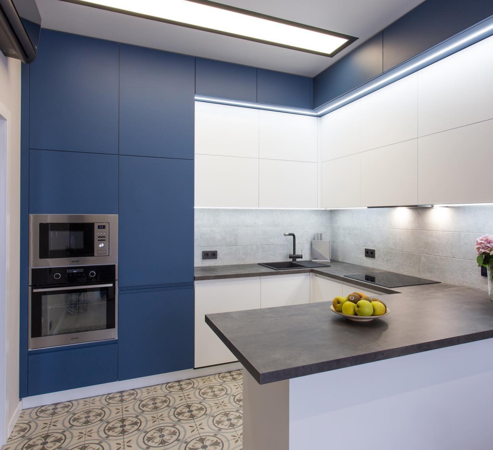 Modern inredning av ett avskilt, mellanstort svart svart l-kök, med en nedsänkt diskho, släta luckor, blå skåp, grått stänkskydd, stänkskydd i stenkakel, rostfria vitvaror, cementgolv, en halv köksö och beiget golv