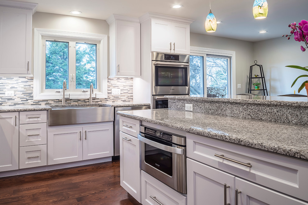Mid-sized transitional u-shaped medium tone wood floor eat-in kitchen photo in Chicago with a farmhouse sink, recessed-panel cabinets, white cabinets, granite countertops, gray backsplash, glass tile backsplash, stainless steel appliances and an island