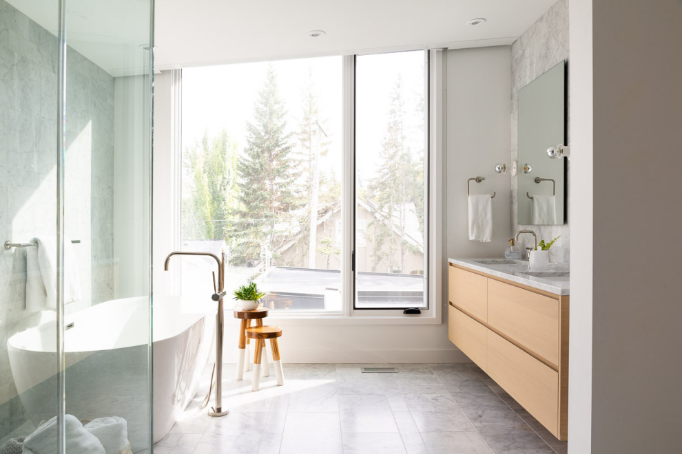 This is an example of a mid-sized contemporary master bathroom in Calgary with light wood cabinets, a freestanding tub, a curbless shower, a one-piece toilet, gray tile, marble, white walls, marble floors, an undermount sink, marble benchtops, grey floor, a hinged shower door, grey benchtops and flat-panel cabinets.