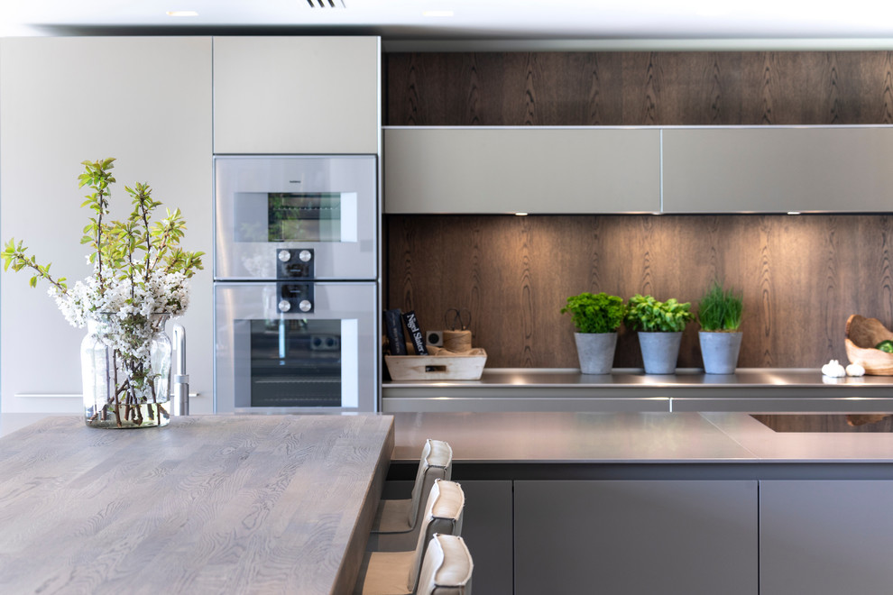 Photo of a large contemporary open plan kitchen in Cheshire with an integrated sink, flat-panel cabinets, medium wood cabinets, stainless steel benchtops, timber splashback, stainless steel appliances, ceramic floors, multiple islands, white floor and green benchtop.