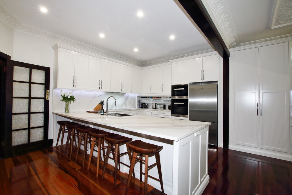 This is an example of a large traditional u-shaped kitchen pantry in Sydney with a single-bowl sink, shaker cabinets, white cabinets, tile benchtops, multi-coloured splashback, porcelain splashback, stainless steel appliances, dark hardwood floors, a peninsula and brown floor.