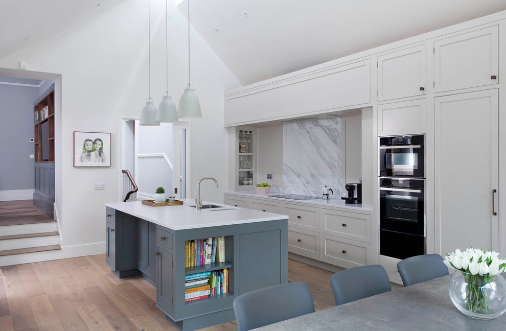 This is an example of a transitional galley eat-in kitchen in Dublin with a double-bowl sink, recessed-panel cabinets, white cabinets, grey splashback, light hardwood floors, with island and beige floor.