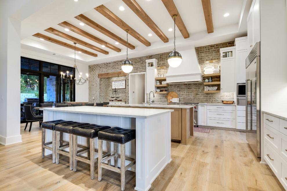 Country eat-in kitchen in Austin with shaker cabinets, white cabinets, brick splashback, stainless steel appliances, light hardwood floors, multiple islands and grey benchtop.