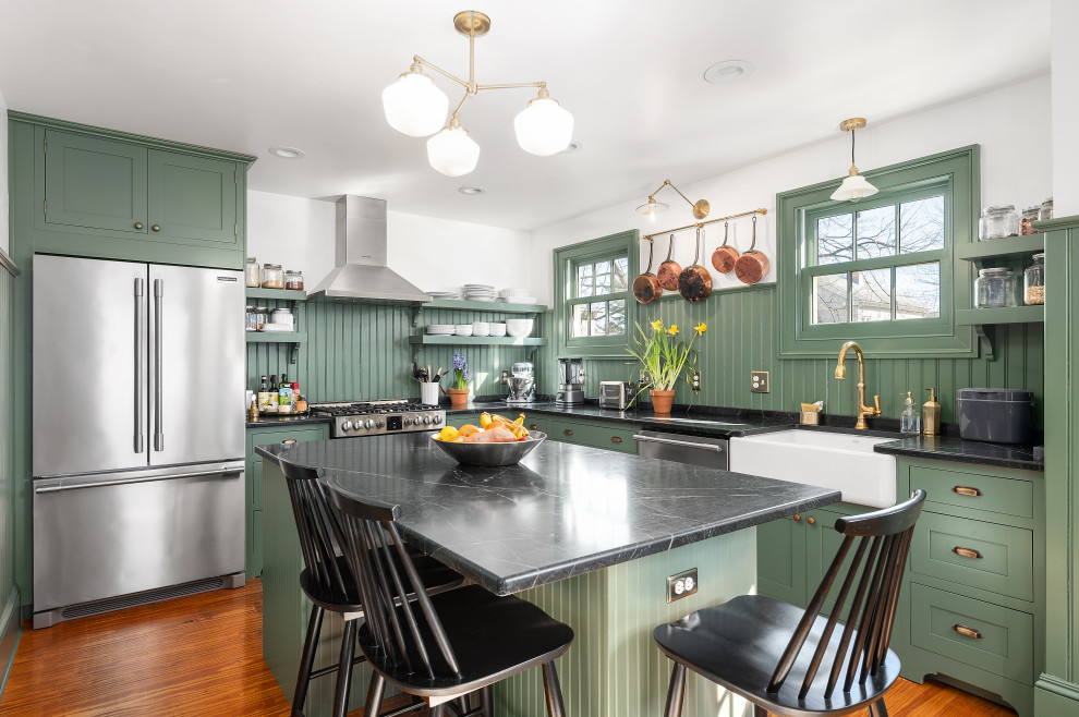 Example of a classic l-shaped medium tone wood floor and brown floor kitchen design in Providence with a farmhouse sink, shaker cabinets, green cabinets, green backsplash, wood backsplash, stainless steel appliances, an island and black countertops
