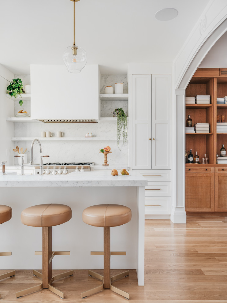 Design ideas for a transitional kitchen in San Francisco with an integrated sink, shaker cabinets, white cabinets, white splashback, stone slab splashback, medium hardwood floors, with island, brown floor and white benchtop.
