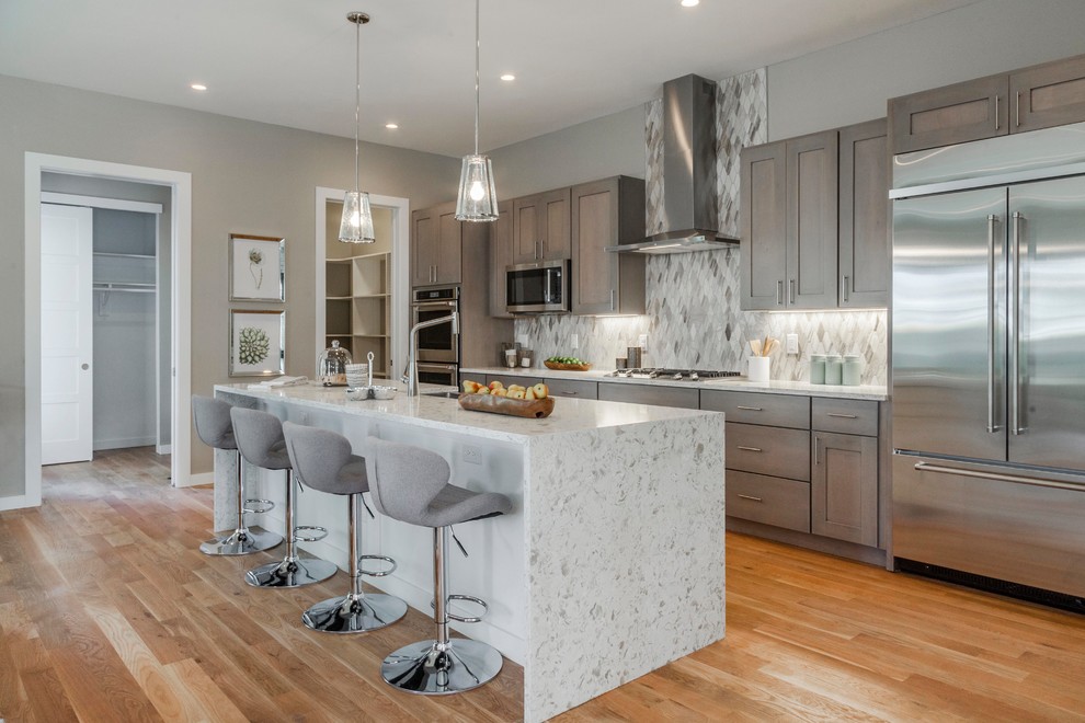 This is an example of a transitional galley kitchen in Denver with shaker cabinets, grey cabinets, grey splashback, stainless steel appliances, medium hardwood floors, with island, brown floor and white benchtop.