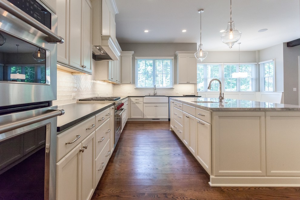 Example of a transitional u-shaped medium tone wood floor and brown floor open concept kitchen design in DC Metro with a farmhouse sink, recessed-panel cabinets, white cabinets, white backsplash, subway tile backsplash, stainless steel appliances, an island and black countertops