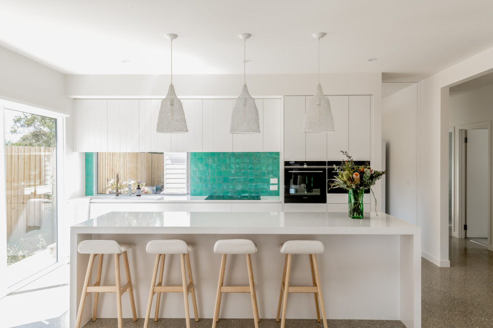 This is an example of a beach style galley kitchen in Geelong with flat-panel cabinets, white cabinets, window splashback, black appliances, concrete floors, with island, grey floor and white benchtop.