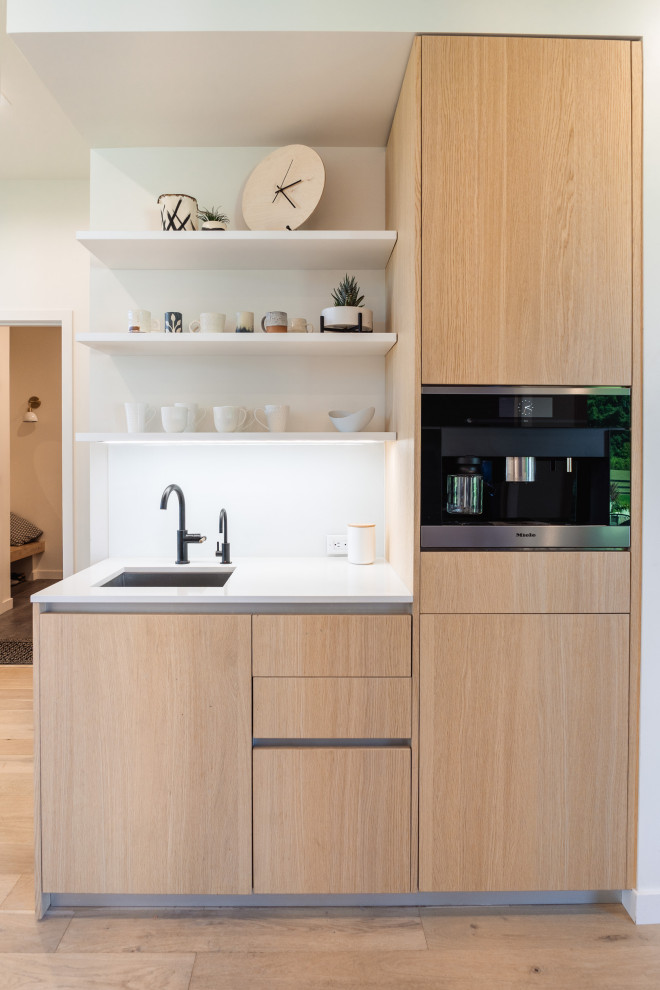 This is an example of an expansive modern single-wall eat-in kitchen in Portland with an undermount sink, flat-panel cabinets, light wood cabinets, quartz benchtops, white splashback, glass sheet splashback, panelled appliances, light hardwood floors, with island, brown floor and white benchtop.
