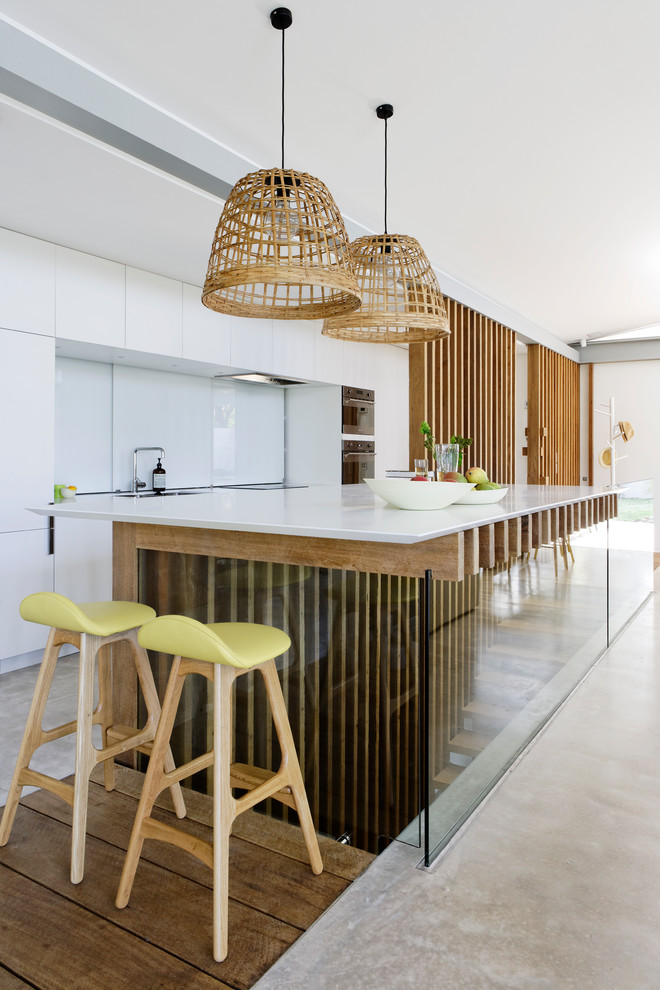 Photo of a mid-sized contemporary galley open plan kitchen in Sydney with a double-bowl sink, beaded inset cabinets, medium wood cabinets, quartz benchtops, white splashback, glass sheet splashback, black appliances, concrete floors, with island, grey floor and white benchtop.