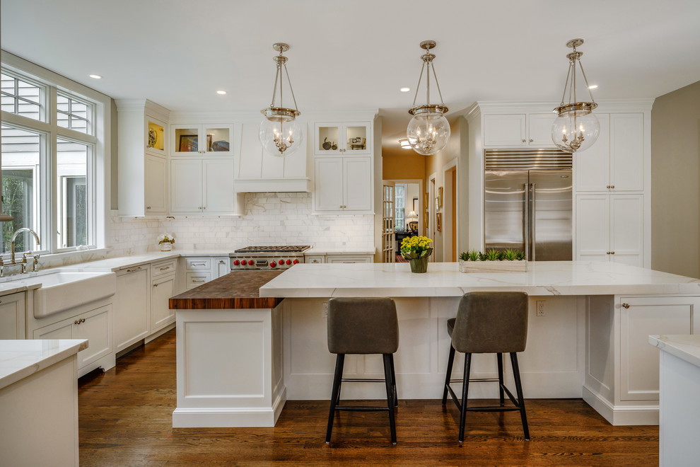 Example of a large transitional u-shaped dark wood floor and brown floor kitchen design in Bridgeport with a farmhouse sink, white cabinets, quartz countertops, white backsplash, marble backsplash, stainless steel appliances, an island, white countertops and recessed-panel cabinets