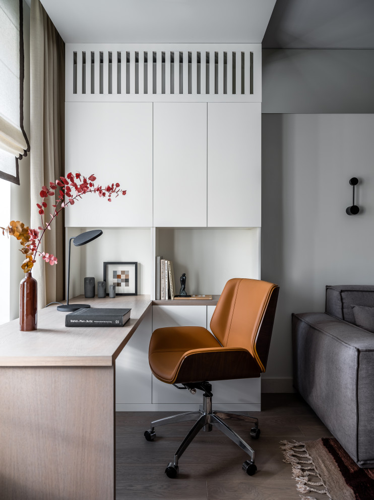 Modelo de despacho actual de tamaño medio sin chimenea con paredes blancas, suelo de madera en tonos medios, escritorio empotrado, suelo gris y bandeja