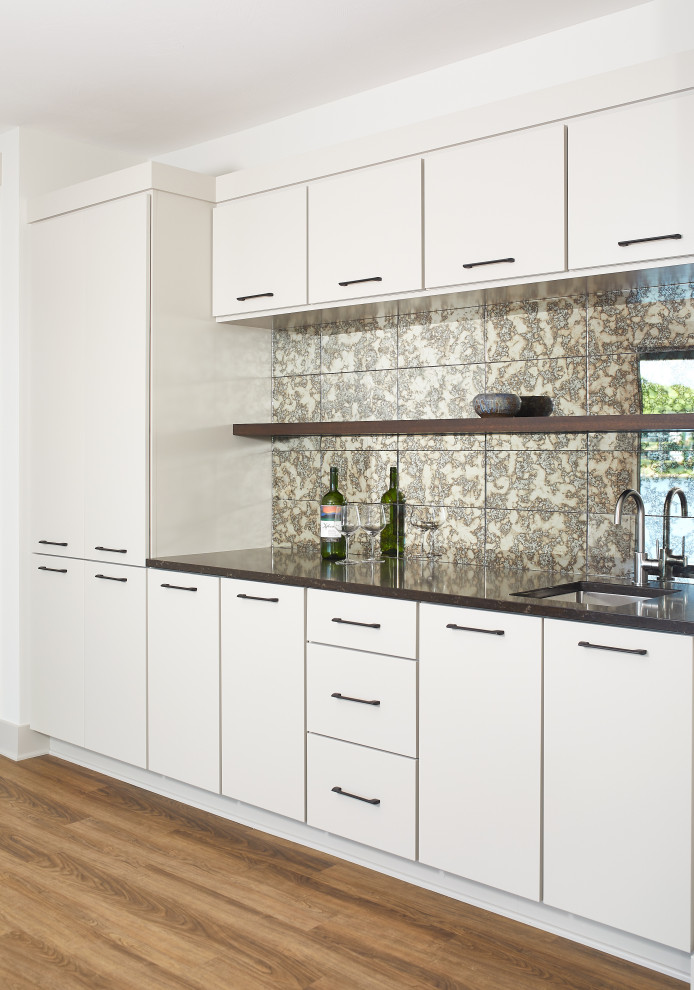 This is an example of a modern single-wall wet bar in Grand Rapids with an undermount sink, flat-panel cabinets, white cabinets, granite benchtops, multi-coloured splashback, mirror splashback, light hardwood floors, brown floor and black benchtop.