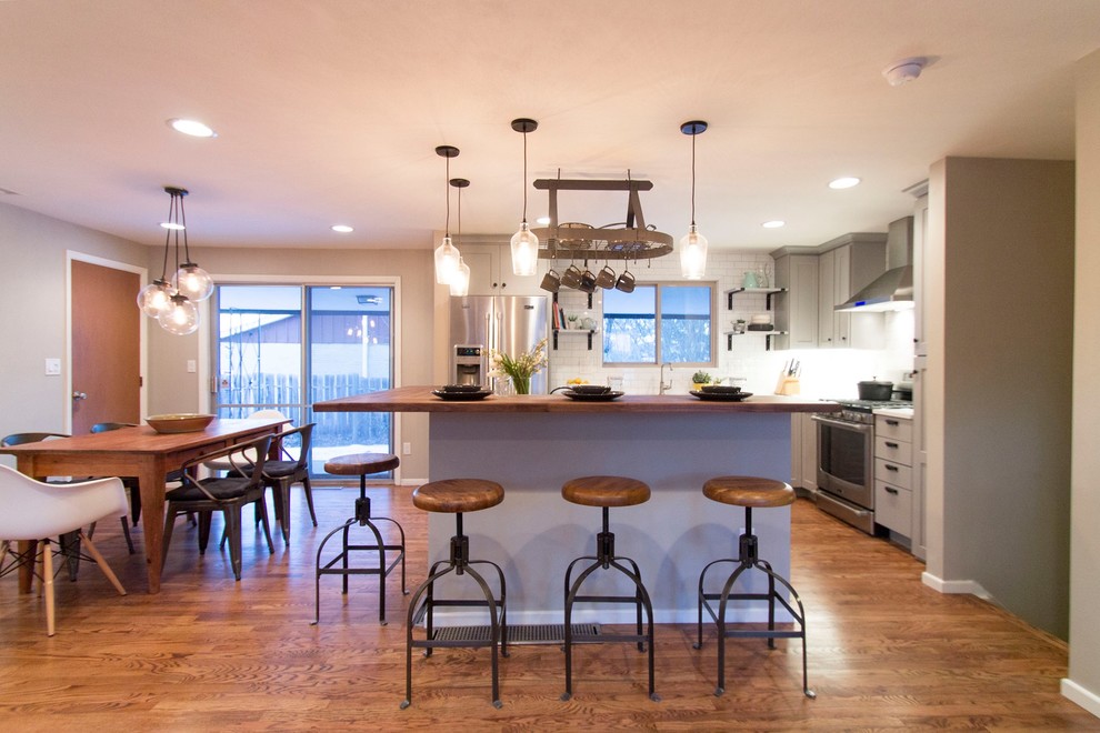 Example of a mid-sized transitional l-shaped medium tone wood floor eat-in kitchen design in Denver with a farmhouse sink, shaker cabinets, gray cabinets, quartz countertops, white backsplash, ceramic backsplash, stainless steel appliances and an island