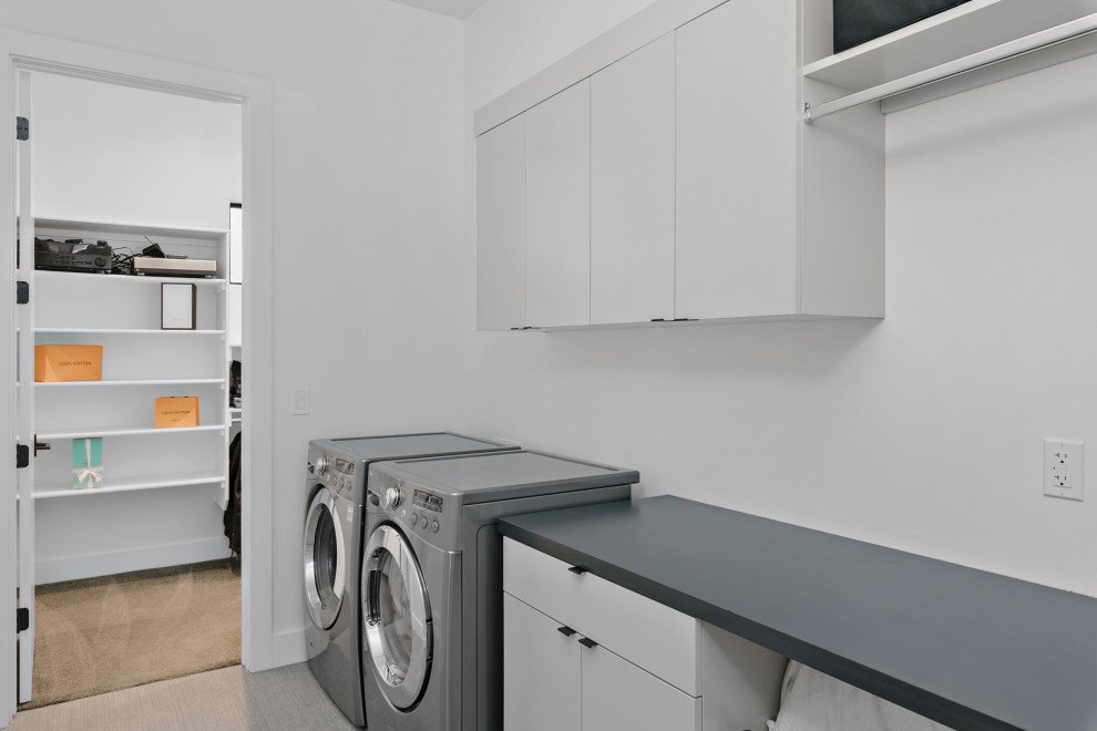 Idéer för att renovera en stor funkis grå linjär grått tvättstuga enbart för tvätt, med släta luckor, vita skåp, laminatbänkskiva, en tvättmaskin och torktumlare bredvid varandra, vitt stänkskydd, vita väggar, heltäckningsmatta och beiget golv
