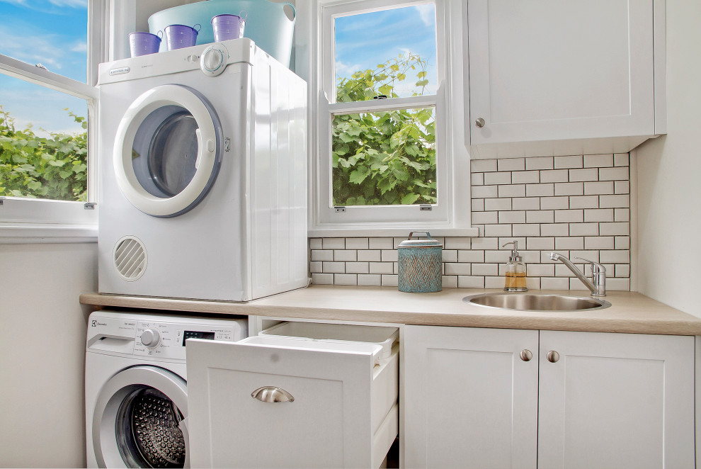 This is an example of a small contemporary laundry room in Sydney with a single-bowl sink, shaker cabinets, white cabinets, laminate benchtops, white splashback, ceramic splashback and a stacked washer and dryer.