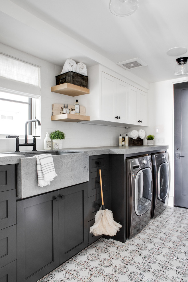 This is an example of a beach style single-wall dedicated laundry room in Los Angeles with a farmhouse sink, shaker cabinets, grey cabinets, white walls, a side-by-side washer and dryer, multi-coloured floor and grey benchtop.