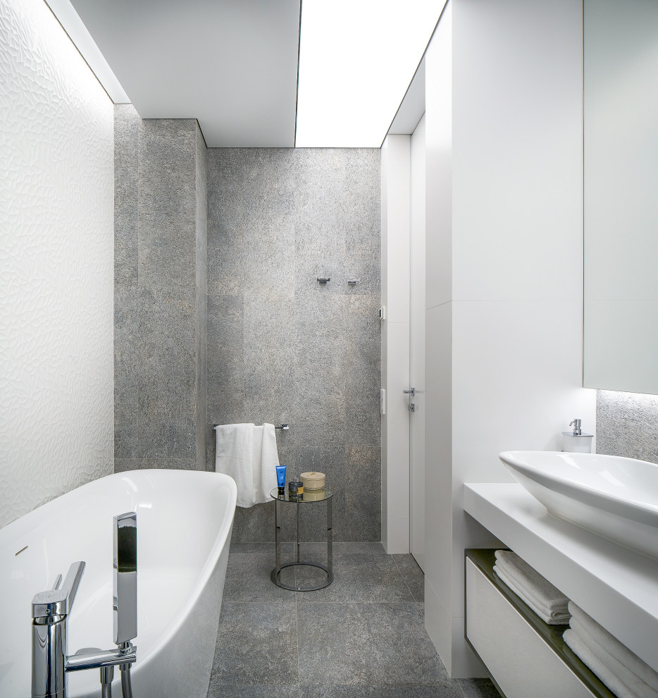 Cette photo montre une salle de bain principale tendance de taille moyenne avec un placard à porte plane, des portes de placard blanches, une baignoire indépendante, WC suspendus, un carrelage blanc, des carreaux de porcelaine, un mur blanc, un sol en carrelage de porcelaine, une vasque, un plan de toilette en surface solide, un sol gris, un plan de toilette blanc, une niche, meuble simple vasque, meuble-lavabo encastré et un plafond décaissé.