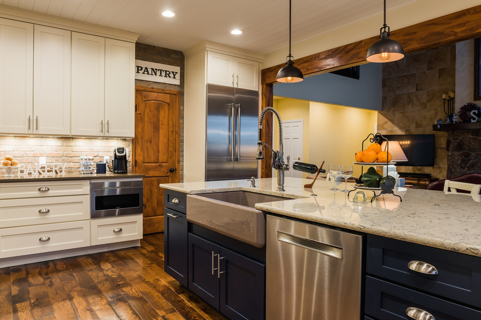 Photo of a mid-sized country eat-in kitchen in Denver with a farmhouse sink, shaker cabinets, white cabinets, quartz benchtops, grey splashback, brick splashback, stainless steel appliances, medium hardwood floors, with island, brown floor and grey benchtop.