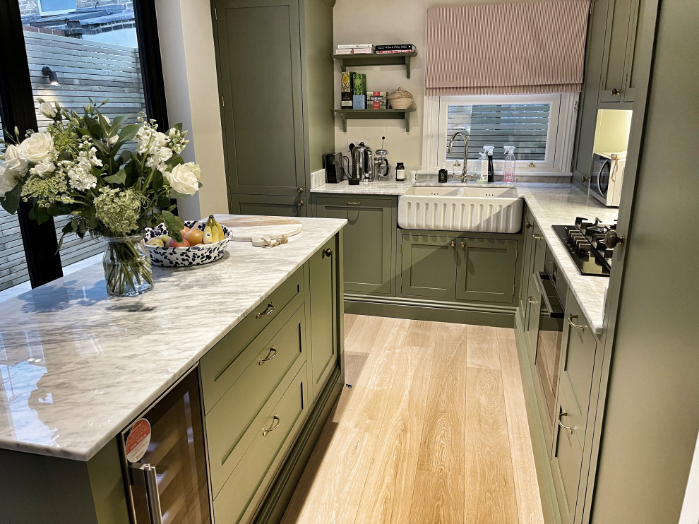 This is an example of a medium sized traditional l-shaped open plan kitchen in London with a belfast sink, shaker cabinets, green cabinets, quartz worktops, white splashback, an island and white worktops.
