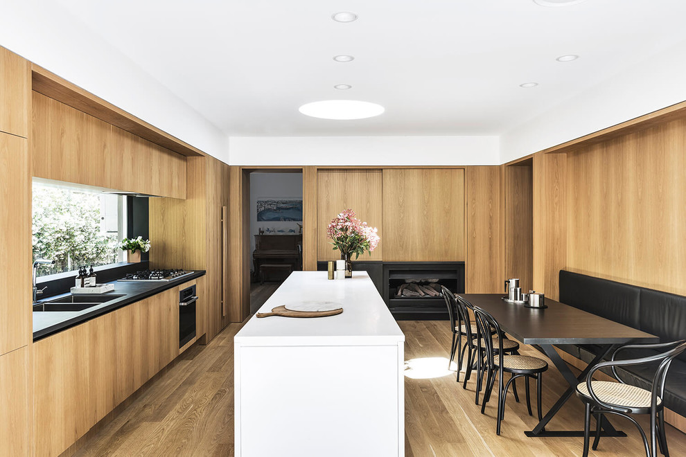 This is an example of a contemporary galley eat-in kitchen in Sydney with a double-bowl sink, flat-panel cabinets, medium wood cabinets, black splashback, window splashback, panelled appliances, medium hardwood floors, with island, brown floor and black benchtop.