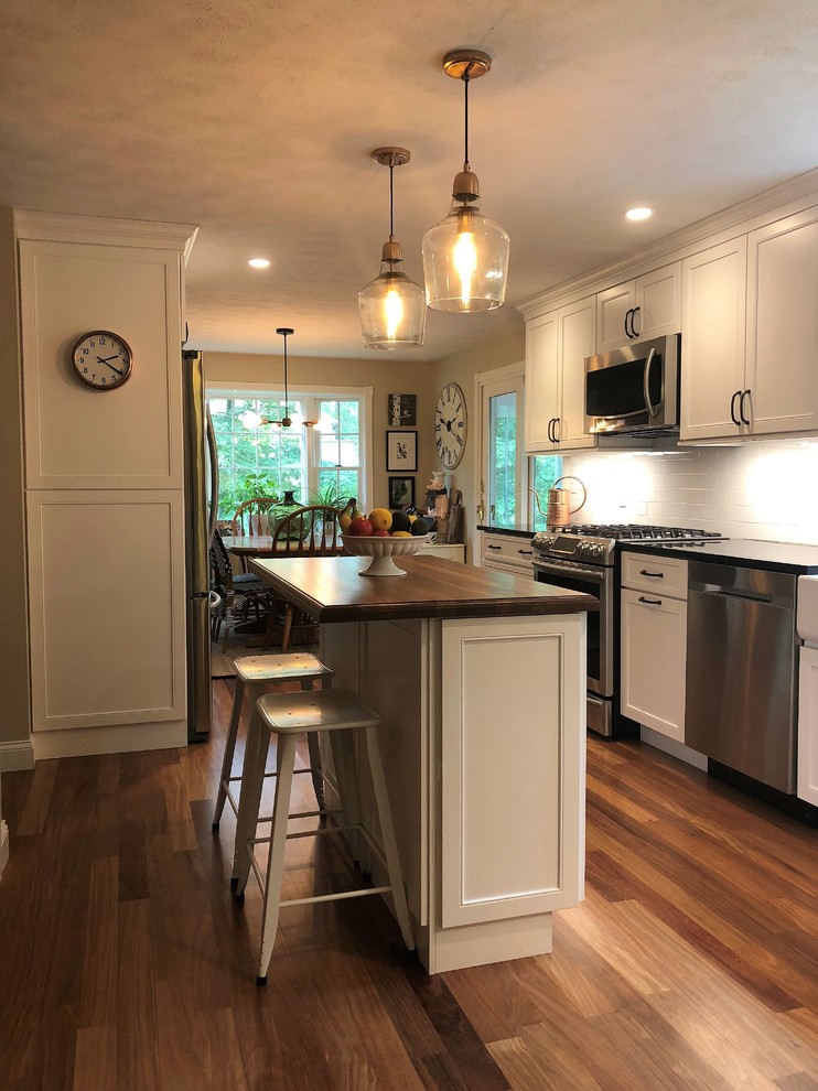 Photo of a mid-sized country l-shaped eat-in kitchen in Boston with a farmhouse sink, shaker cabinets, white cabinets, wood benchtops, white splashback, porcelain splashback, stainless steel appliances, medium hardwood floors, with island, multi-coloured floor and brown benchtop.