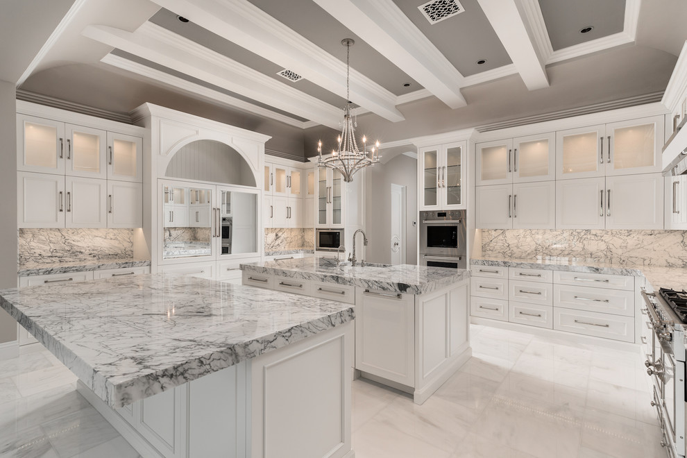 This is an example of an expansive modern u-shaped kitchen in Phoenix with a farmhouse sink, glass-front cabinets, white cabinets, marble benchtops, marble splashback, stainless steel appliances, marble floors, multiple islands, grey floor and white benchtop.