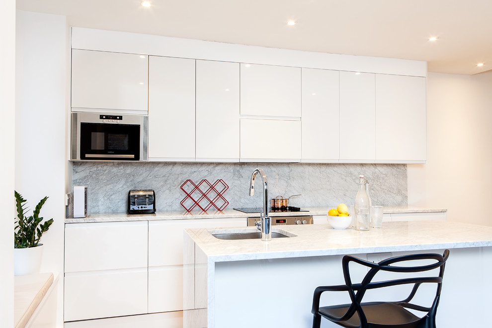 Photo of a contemporary galley kitchen in London with an undermount sink, flat-panel cabinets, white cabinets, grey splashback, white appliances and with island.