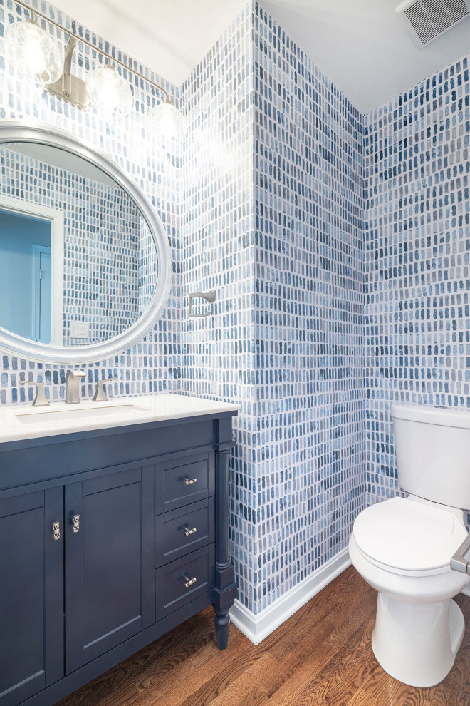 Design ideas for a small beach style powder room in DC Metro with furniture-like cabinets, blue cabinets, a one-piece toilet, blue tile, multi-coloured walls, medium hardwood floors, an undermount sink, engineered quartz benchtops, brown floor, white benchtops, a freestanding vanity and wallpaper.