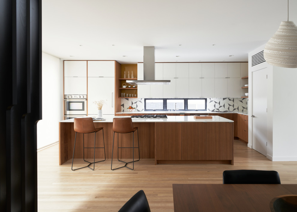Photo of a modern l-shaped eat-in kitchen in Seattle with a double-bowl sink, flat-panel cabinets, medium wood cabinets, quartz benchtops, multi-coloured splashback, cement tile splashback, stainless steel appliances, light hardwood floors, with island and white benchtop.