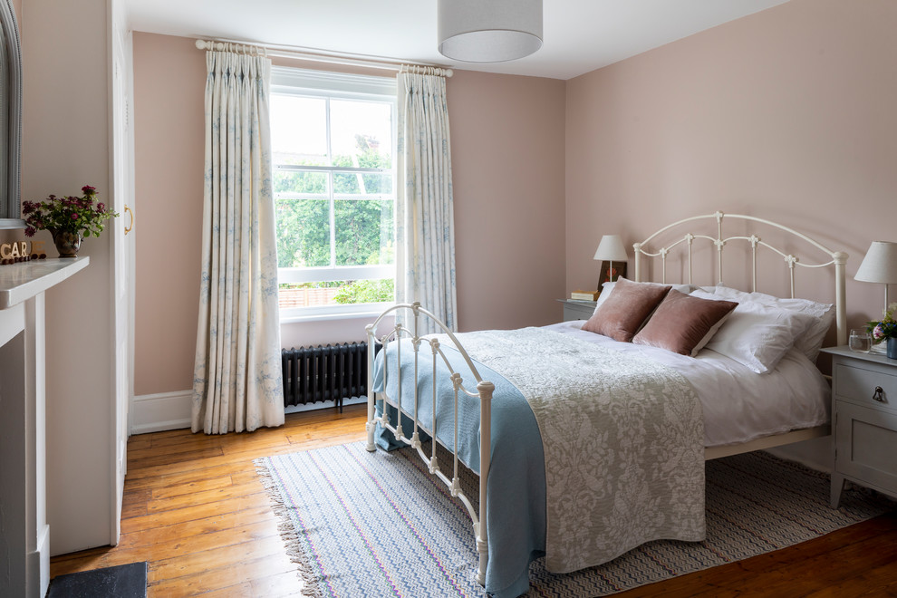Mid-sized traditional guest bedroom in London with pink walls, light hardwood floors, a standard fireplace and beige floor.