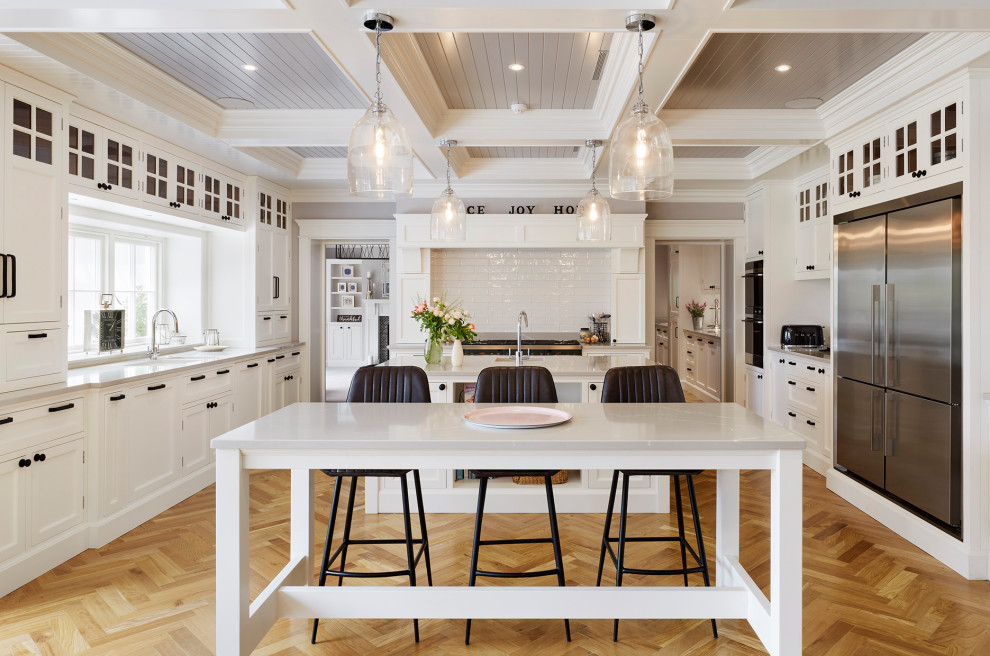 Photo of a beach style u-shaped separate kitchen in Boston with an undermount sink, glass-front cabinets, white cabinets, white splashback, subway tile splashback, stainless steel appliances, light hardwood floors, multiple islands, beige floor and grey benchtop.