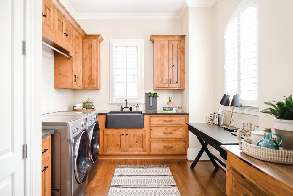 Inspiration for a transitional l-shaped utility room in Charlotte with a farmhouse sink, shaker cabinets, medium wood cabinets, brown walls, a side-by-side washer and dryer, grey benchtop and medium hardwood floors.