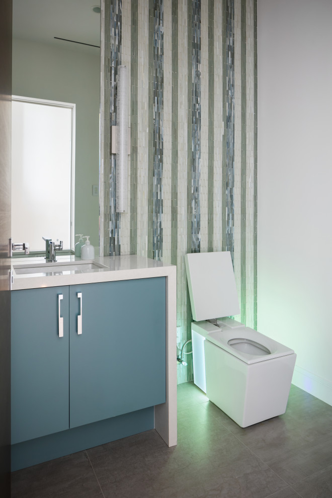 На фото: туалет среднего размера в современном стиле с плоскими фасадами, зелеными фасадами, унитазом-моноблоком, разноцветной плиткой, стеклянной плиткой, разноцветными стенами, полом из керамогранита, врезной раковиной, столешницей из искусственного кварца, серым полом, белой столешницей и встроенной тумбой с