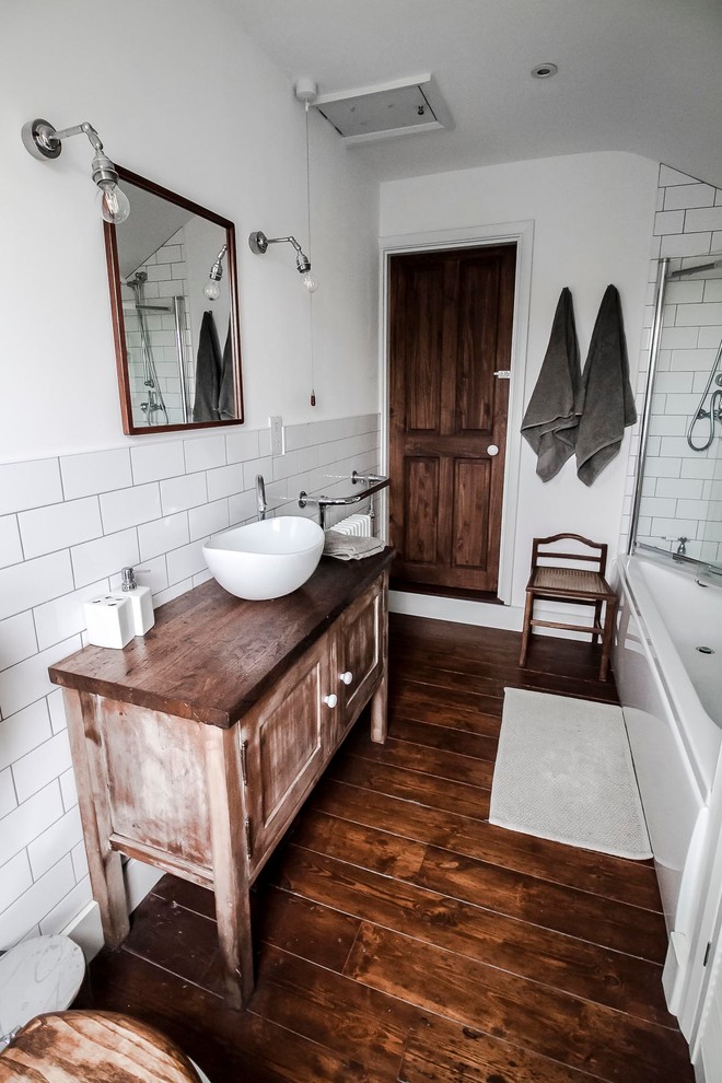 Mid-sized traditional kids bathroom in London with shaker cabinets, brown cabinets, a drop-in tub, a shower/bathtub combo, white tile, subway tile, white walls, dark hardwood floors, a vessel sink, wood benchtops, brown floor, a sliding shower screen and a one-piece toilet.