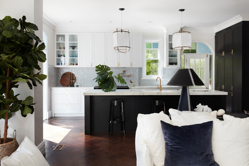Photo of a transitional l-shaped open plan kitchen in Sydney with a farmhouse sink, shaker cabinets, grey splashback, black appliances, dark hardwood floors, with island and white benchtop.