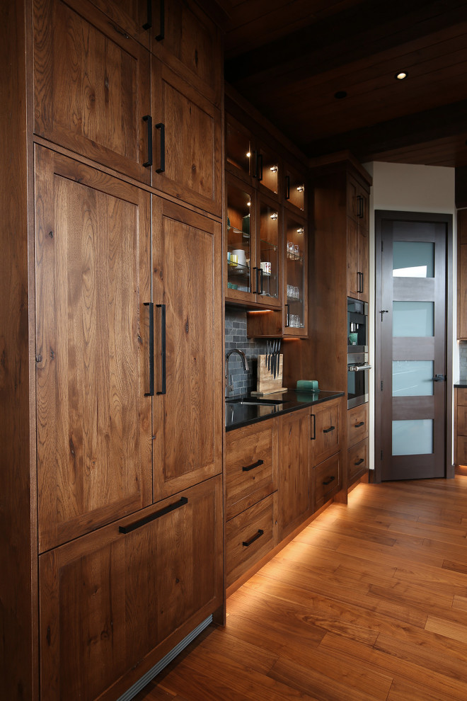 Example of a large mountain style u-shaped medium tone wood floor, brown floor and exposed beam open concept kitchen design with an undermount sink, shaker cabinets, medium tone wood cabinets, quartz countertops, gray backsplash, slate backsplash, stainless steel appliances, two islands and black countertops