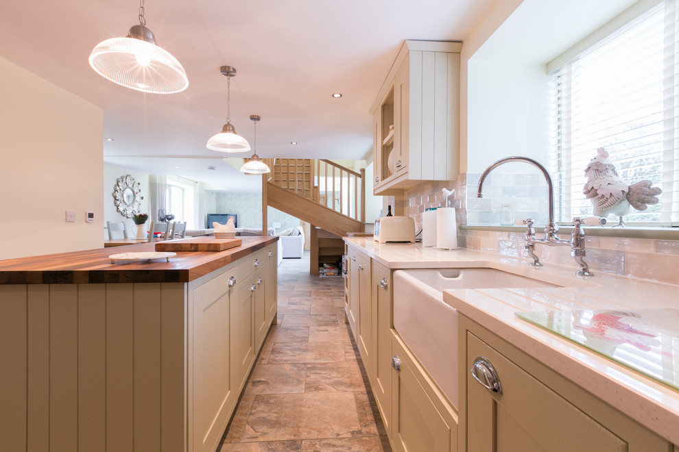 Photo of a medium sized country l-shaped kitchen/diner with a belfast sink, shaker cabinets, green cabinets, wood worktops, multi-coloured splashback, stone tiled splashback, stainless steel appliances, ceramic flooring and an island.