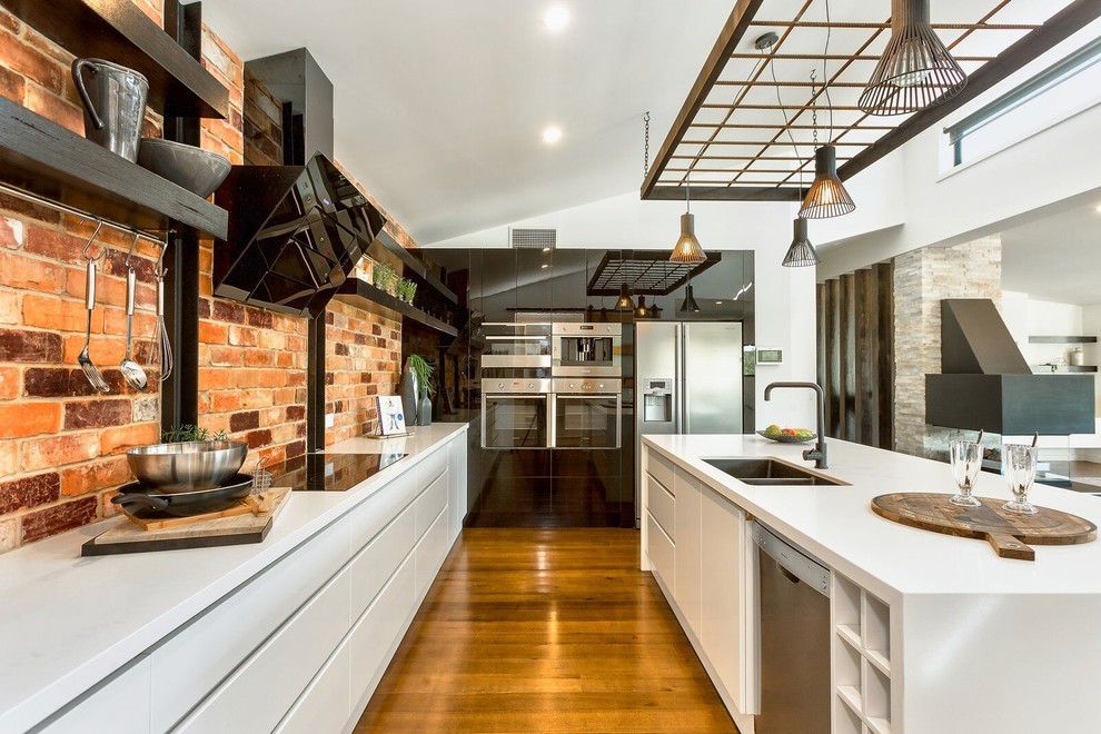 Large industrial l-shaped eat-in kitchen in Other with an undermount sink, black cabinets, quartz benchtops, red splashback, brick splashback, stainless steel appliances, with island, brown floor and white benchtop.