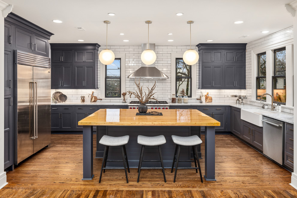 Design ideas for a transitional u-shaped kitchen in Nashville with a farmhouse sink, recessed-panel cabinets, black cabinets, white splashback, subway tile splashback, stainless steel appliances, medium hardwood floors, with island, brown floor and grey benchtop.