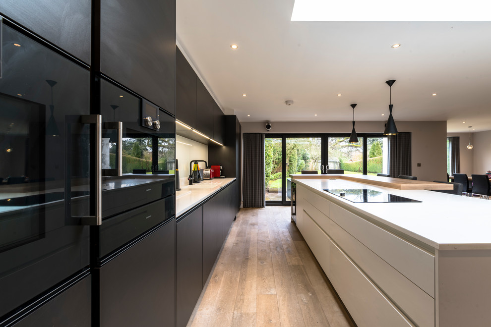 Large contemporary l-shaped open plan kitchen in Cheshire with a drop-in sink, flat-panel cabinets, solid surface benchtops, white splashback, glass sheet splashback, black appliances, light hardwood floors and with island.