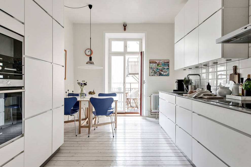 This is an example of a mid-sized scandinavian galley eat-in kitchen in Gothenburg with flat-panel cabinets, white cabinets, stainless steel benchtops, porcelain splashback, panelled appliances, light hardwood floors, no island, beige floor and white splashback.
