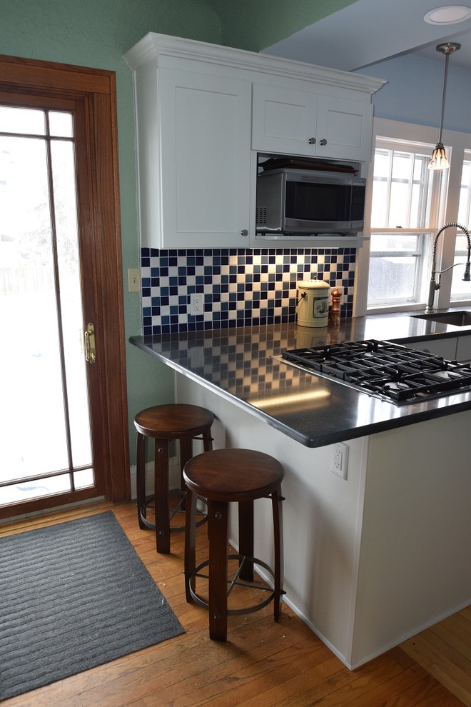 Example of a small transitional l-shaped light wood floor kitchen design with an undermount sink, shaker cabinets, white cabinets, quartz countertops, multicolored backsplash, ceramic backsplash, stainless steel appliances and a peninsula