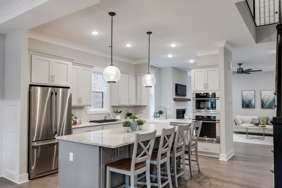 Mid-sized traditional l-shaped eat-in kitchen in Richmond with a farmhouse sink, shaker cabinets, white cabinets, quartzite benchtops, white splashback, subway tile splashback, stainless steel appliances, medium hardwood floors, with island, brown floor and white benchtop.