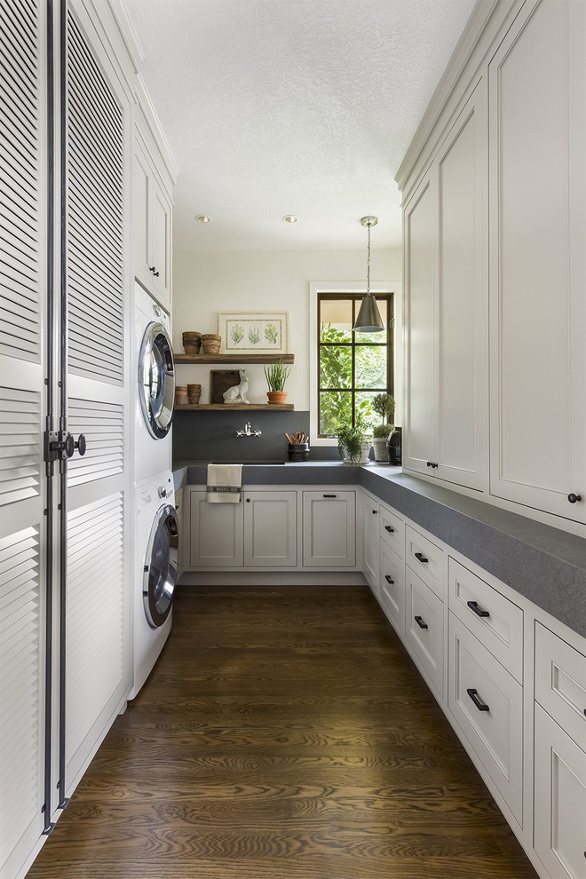 Photo of a traditional u-shaped laundry room in Portland with an undermount sink, recessed-panel cabinets, white cabinets, white walls, dark hardwood floors, a stacked washer and dryer and grey benchtop.