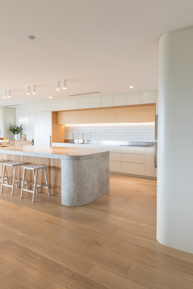 Foto på ett stort funkis grå kök, med en integrerad diskho, vita skåp, bänkskiva i kalksten, vitt stänkskydd, stänkskydd i cementkakel, integrerade vitvaror, ljust trägolv, en köksö och beiget golv