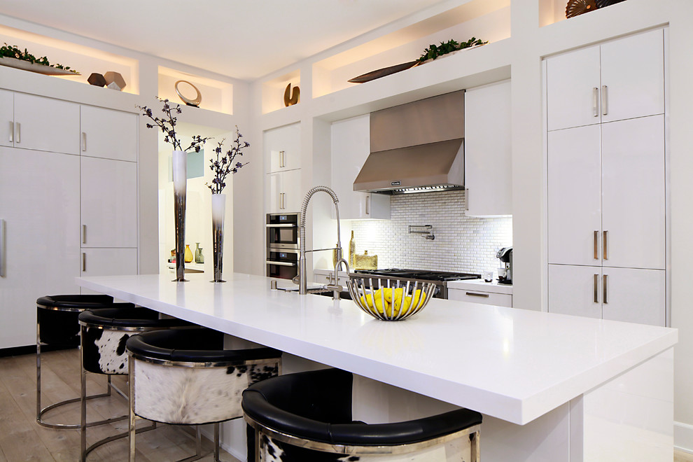 Inspiration for a mid-sized contemporary l-shaped open plan kitchen in Los Angeles with a drop-in sink, flat-panel cabinets, white cabinets, quartzite benchtops, white splashback, glass tile splashback, stainless steel appliances, light hardwood floors, with island, beige floor and white benchtop.