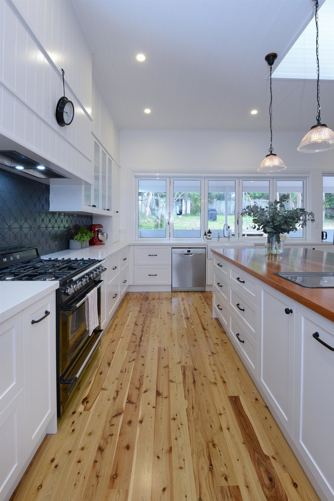 Inspiration for a large traditional u-shaped open plan kitchen in Sydney with a drop-in sink, recessed-panel cabinets, white cabinets, wood benchtops, black splashback, ceramic splashback, black appliances, light hardwood floors, with island, brown floor and grey benchtop.