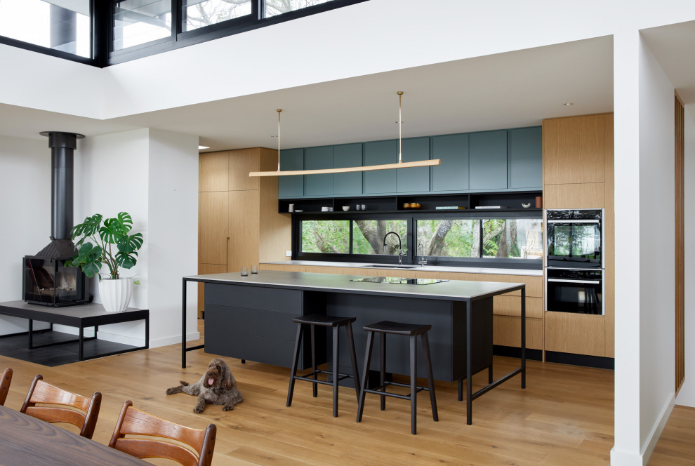 Photo of a large contemporary single-wall eat-in kitchen in Melbourne with an undermount sink, flat-panel cabinets, light wood cabinets, solid surface benchtops, black appliances, medium hardwood floors, with island and white benchtop.